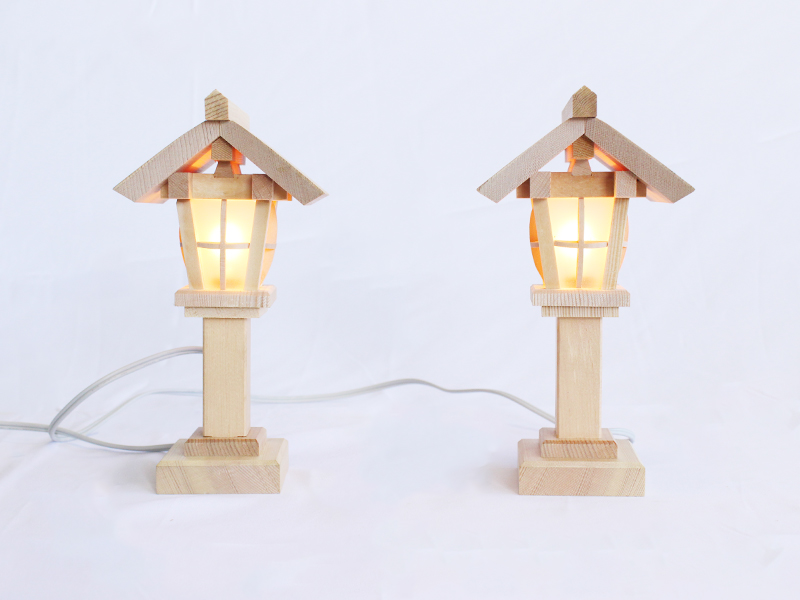 A pair of wooden lamps with roof-like tops on poles, akin to a miniature streetlamp. There is a white electrical cord extending from both.
