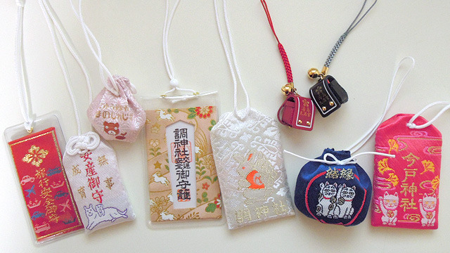 A variety of omamori laid on a white surface. Most are made of embroided cloth and some are encased in vinyl. Some are shaped like rectangles, while others are shaped like pouches or children's backpacks. All bear writing indicating the shrine of origin and their intended blessing.
